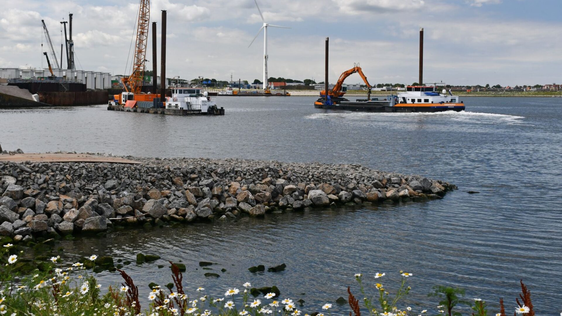 zicht op kraanschepen op water met op de voorgrond een stenen breukdam en witte bloemetjes