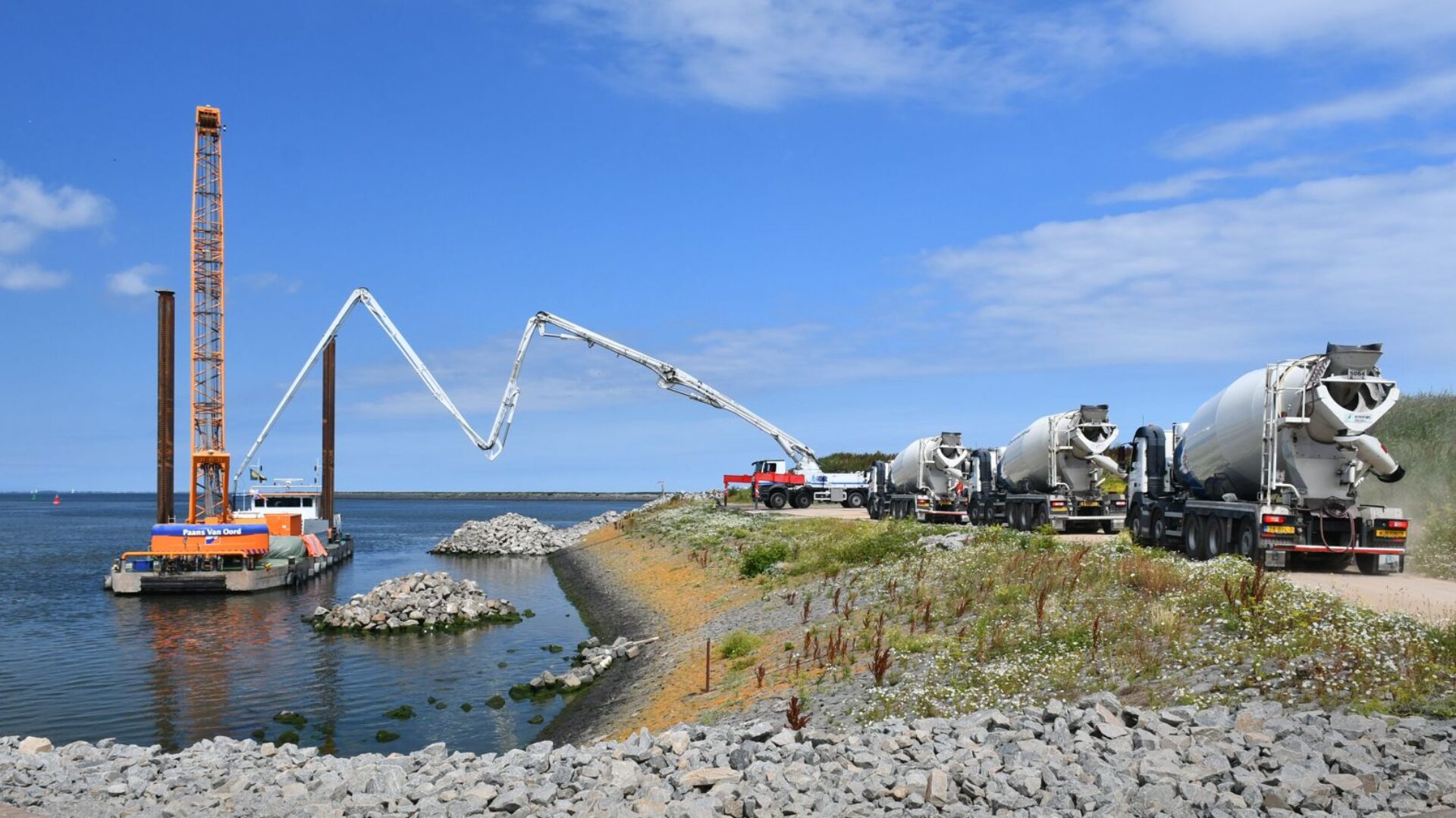 blauwe lucht met links water met een kraanschip en rechts een oever met betonmolens. Het beton uit een betonmolen wordt via een pijp geleid naar het schip.