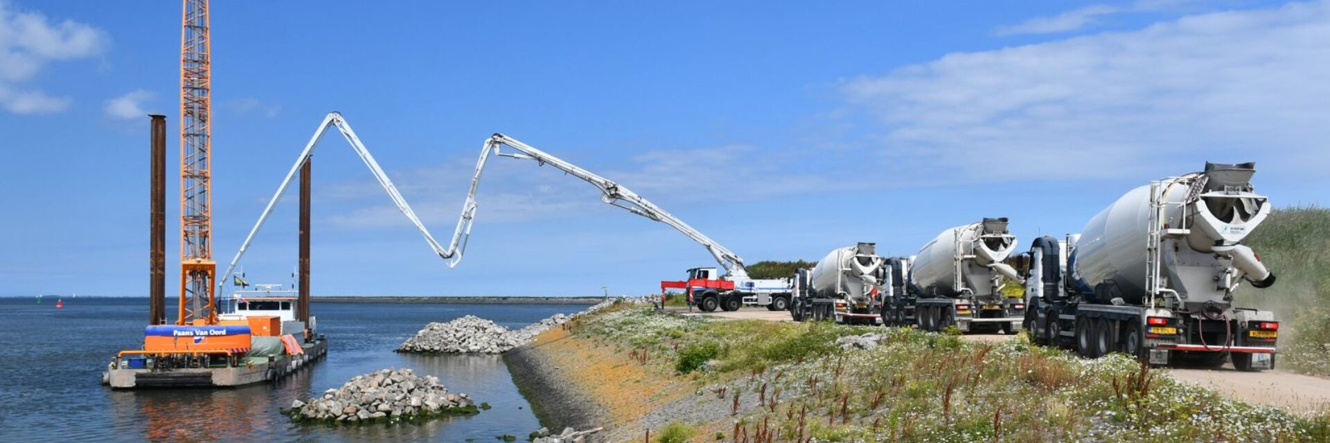 blauwe lucht met links water met een kraanschip en rechts een oever met betonmolens. Het beton uit een betonmolen wordt via een pijp geleid naar het schip.