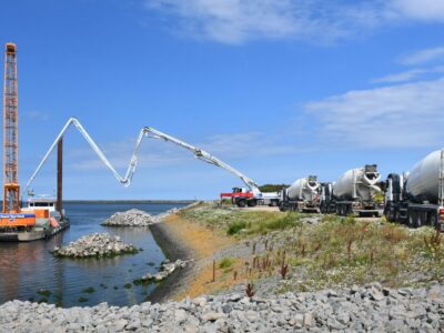 blauwe lucht met links water met een kraanschip en rechts een oever met betonmolens. Het beton uit een betonmolen wordt via een pijp geleid naar het schip.