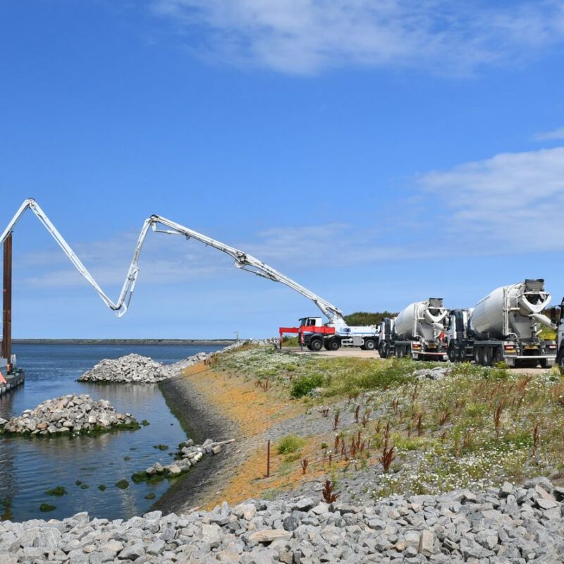 blauwe lucht met links water met een kraanschip en rechts een oever met betonmolens. Het beton uit een betonmolen wordt via een pijp geleid naar het schip.