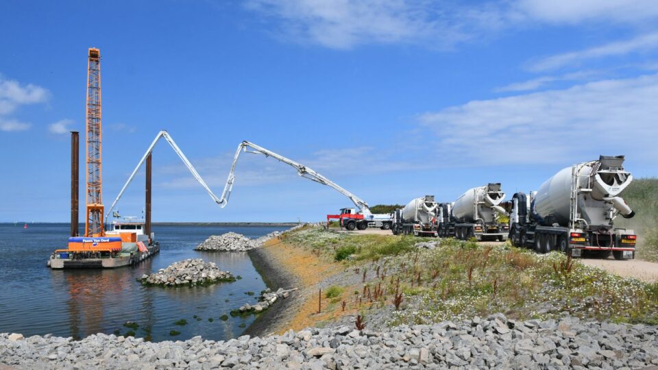 blauwe lucht met links water met een kraanschip en rechts een oever met betonmolens. Het beton uit een betonmolen wordt via een pijp geleid naar het schip.