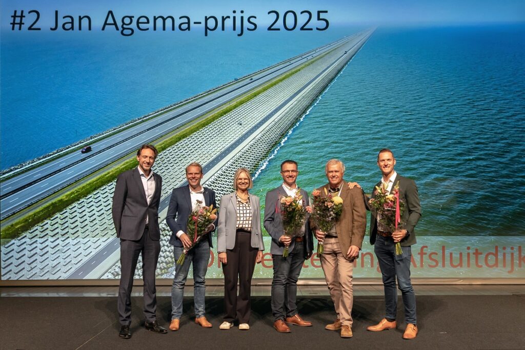 Je ziet 6 mensen staan met bloemen voor een grote wand met een luchtfoto van de dijkversterking van de Afsluitdijk.