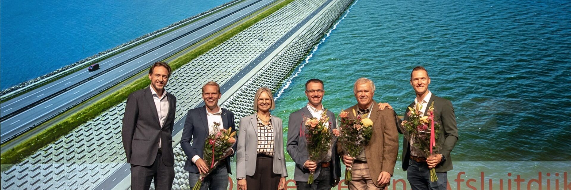 Je ziet 6 mensen staan met bloemen voor een grote wand met een luchtfoto van de dijkversterking van de Afsluitdijk.