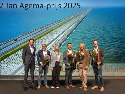 Je ziet 6 mensen staan met bloemen voor een grote wand met een luchtfoto van de dijkversterking van de Afsluitdijk.