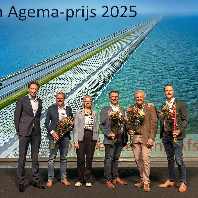 Je ziet 6 mensen staan met bloemen voor een grote wand met een luchtfoto van de dijkversterking van de Afsluitdijk.