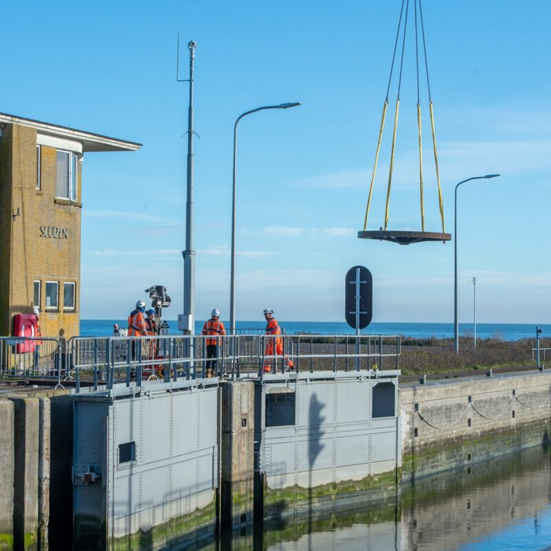 We zien dat een tandwiel in de lucht gehesen is bij een sluiscomplex.