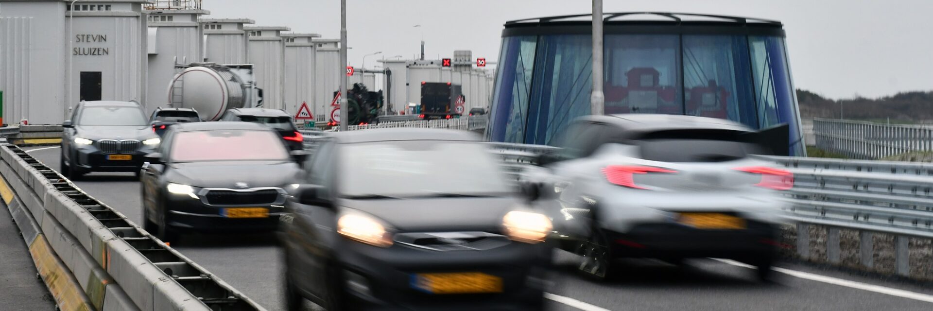 we zien auto's rijden over de rijbaan met op de achtergrond witte gebouwtjes en een koepel die typerend zijn voor de Afsluitdijk.